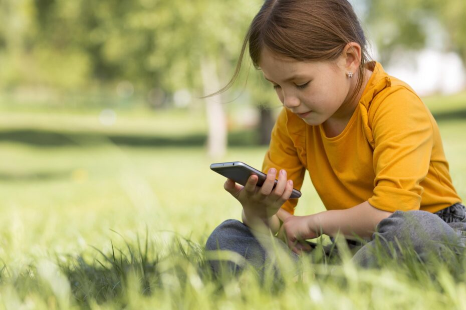Imagem de Freepik mostra criança sentada na grama olhando para um celular